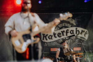 Katerfahrt live Auftritt der Band Katerfahrt beim Elbenwald Festival, mit einem fokussierten Bassisten im Vordergrund und einem Schlagzeuger im Hintergrund, der traditionell gekleidet ist. Bunte Lichter und ein lebhaftes Festival-Setting schaffen eine energiegeladene Atmosphäre.