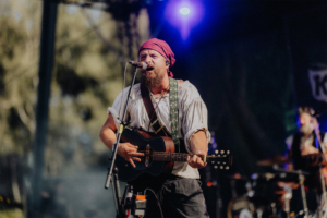 Katerfahrt live Ein Musiker mit rotem Kopftuch spielt auf der Bühne des Elbenwald Festivals eine Gitarre. Er trägt ein historisches Outfit und singt leidenschaftlich ins Mikrofon, während die bunten Lichter der Veranstaltung die Atmosphäre beleuchten.