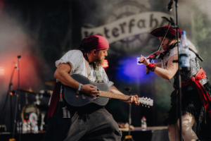 Katerfahrt live Zwei Musiker auf der Bühne des Elbenwald Festivals, ein Mann mit Gitarre und eine Frau mit Geige, beide in fantasievoller Kleidung. Bunte Lichter und eine lebendige Atmosphäre schaffen ein einzigartiges Festivalerlebnis für Fans der Popkultur.