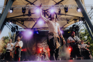 Katerfahrt live Ein Musiker in Piratenkostüm steht auf der Bühne des Elbenwald Festivals und hält ein Mikrofon hoch, während eine Band im Hintergrund spielt. Bunte Lichter und eine begeisterte Menge schaffen eine lebendige Festivalatmosphäre.