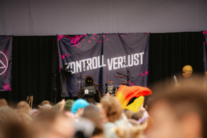 Kontrollverlust Bühne beim Elbenwald Festival mit dem Banner "Kontroll Verlust". Im Vordergrund sind Zuschauer zu sehen, während eine Band mit futuristischen Kostümen auftritt. Die Atmosphäre ist lebhaft und bunt, ideal für Fans von Musik und Nerd-Kultur.