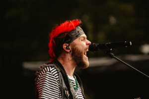 Kontrollverlust Ein Sänger mit rotem Haar und Bandana, der leidenschaftlich in ein Mikrofon am Elbenwald Festival performt. Er trägt ein gestreiftes Shirt und eine Weste, während er von einer lebhaften Festivalatmosphäre umgeben ist.
