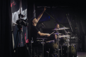 Kontrollverlust Ein Schlagzeuger spielt leidenschaftlich auf der Bühne des Elbenwald Festivals, während ein Kamerateam die Performance aufnimmt. Die Atmosphäre ist lebhaft und energiegeladen, mit einer Mischung aus Musik und nerdiger Kultur im Hintergrund.