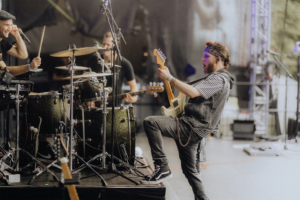 Kontrollverlust Musiker auf der Bühne beim Elbenwald Festival, der mit Leidenschaft Gitarre spielt. Im Hintergrund sind Schlagzeuger und weitere Bandmitglieder zu sehen, die die energiegeladene Atmosphäre des Festivals unterstreichen.