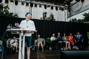Live Quiz Ein Moderator steht an einem Tisch mit Laptop und Getränk auf der Bühne des Elbenwald Festivals, während eine Gruppe von Cosplayern im Hintergrund sitzt. Der Veranstaltungsort ist mit Scheinwerfern und Pflanzen dekoriert, die eine lebendige Atmosphäre schaffen.