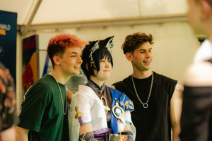Meet and Greet DarkVictory Drei junge Menschen posieren lächelnd beim Elbenwald Festival, während einer im Cosplay-Kostüm mit Katzenohren und Medaillen in der Mitte steht. Die Szene strahlt Freude und Gemeinschaftsgefühl aus, typisch für das Festival, das Musik und Nerdkultur vereint.