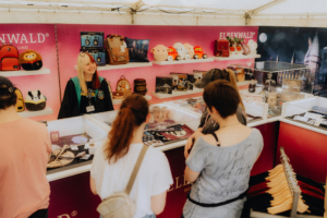 Besucher des Elbenwald Festivals stehen an einem Verkaufsstand mit Merchandise-Artikeln, darunter Taschen, Plüschtiere und Fanartikel. Eine Verkäuferin lächelt, während sie die Produkte präsentiert, umgeben von einem bunten und einladenden Standdesign.