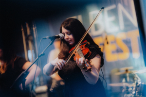 Mardi Musikerin spielt Geige auf der Bühne des Elbenwald Festivals, umgeben von einer lebhaften Atmosphäre. Im Hintergrund sind verschwommen weitere Bandmitglieder und das Festival-Logo sichtbar, das die besondere Verbindung von Musik und Nerd-Kultur betont.