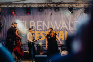 Mardi Musiker auf der Bühne des Elbenwald Festivals, einem Festival, das Musik und Nerdkultur kombiniert. Die Sängerin mit Mikrofon und die Bandmitglieder, darunter ein Kontrabassist und ein Gitarrist, sorgen für eine lebendige Atmosphäre.