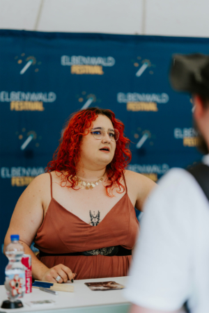 Viiilicious Eine Person mit roten Locken und Brille steht am Tisch des Elbenwald Festivals, umgeben von Merchandise und Fans. Im Hintergrund ist das Festival-Logo sichtbar. Die Atmosphäre ist lebhaft und freundlich, ideal für Nerd- und Musikfans.