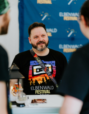 eosAndy Ein lächelnder Mann trägt ein T-Shirt mit dem Logo des Elbenwald Festivals. Er steht an einem Tisch, umgeben von Dekorationen, während er mit einem Besucher spricht. Im Hintergrund ist ein Banner des Festivals sichtbar.