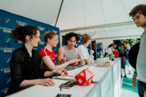 Meet and Greet The Livelines Besucher am Elbenwald Festival stehen an einem Informationsstand, wo sie Merchandise und Spielmaterialien erhalten können. Im Hintergrund sind weitere Festivalteilnehmer und eine festliche Atmosphäre zu sehen.