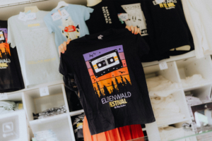 EWF25 Merch Schwarzes T-Shirt mit buntem Elbenwald Festival-Design 2025, das eine Kassette und eine Silhouette von Bäumen zeigt. Im Hintergrund sind weitere T-Shirts im Verkaufsstand zu sehen. Perfekte Merchandise-Option für Festivalbesucher und Nerd-Fans.