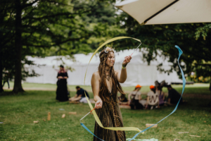 Mitmach-Zirkus Eine Frau mit geflochtenem Haar und Blumenkranz tanzt auf dem Elbenwald Festival, während sie bunte Bänder schwingt. Im Hintergrund sind Festivalbesucher und Zelte zu sehen, die die lebendige Atmosphäre der Veranstaltung unterstreichen.