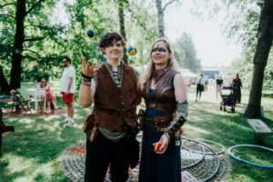 Workshop Mitmachzirkus Zwei Personen in fantasievoller Kleidung jonglieren mit Bällen auf dem Elbenwald Festival. Im Hintergrund sind andere Festivalbesucher und Spielgeräte sichtbar, die die lebendige Atmosphäre des Events unterstreichen.