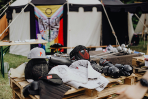 Ein Verkaufsstand auf dem Elbenwald Festival zeigt eine Vielzahl von Cosplay-Ausrüstungen, darunter Helme, Schutzkleidung und Accessoires, vor einem bunten Banner im Hintergrund. Ideal für Fans von Musik und Nerd-Kultur.