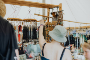 Artist Alley Eine Person mit einem grünen Hut betrachtet Schmuck und Accessoires an einem Stand auf dem Elbenwald Festival, umgeben von bunten Halsketten und Anhängern. Der Stand bietet eine Vielzahl von nerdigen und kreativen Produkten für Festivalbesucher.