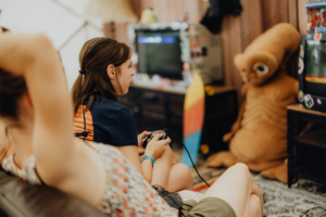 Gamesroom by Nerdistan Eine junge Frau spielt Videospiele auf einem Sofa beim Elbenwald Festival. Im Hintergrund sind ein Fernseher und ein auffälliges, braunes Maskottchen zu sehen. Die Atmosphäre ist entspannt und kreativ, ideal für Nerds und Fans.