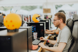 Gamesroom by Nerdistan Ein Teilnehmer am Elbenwald Festival sitzt an einem Computer, umgeben von bunten Dekorationen wie einer Dragon Ball- und Pac-Man-Figur. Die Atmosphäre kombiniert Gaming und kreative Kultur, während er konzentriert an seinem Spiel arbeitet.