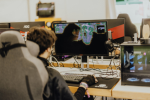 Gamesroom by Nerdistan Ein Teilnehmer des Elbenwald Festivals sitzt an einem Gaming-PC, spielt ein Videospiel auf einem großen Monitor und trägt ein Headset. Im Hintergrund sind weitere Computer und Gaming-Ausrüstung sichtbar, die die nerdige Atmosphäre des Festivals unterstreichen.