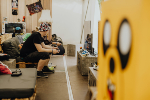 Gamesroom by Nerdistan Ein Besucher spielt Videospiele auf einer Konsole in einer gemütlichen Lounge-Ecke des Elbenwald Festivals. Im Hintergrund sind Figuren und Merchandise aus der Nerdkultur zu sehen, die die entspannte Atmosphäre der Veranstaltung unterstreichen.