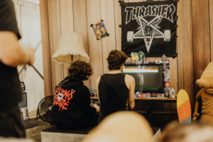 Gamesroom by Nerdistan Zwei Besucher des Elbenwald Festivals sitzen vor einem Retro-Fernseher, auf dem ein Videospiel läuft. Der Raum ist mit einer Thrasher-Flagge, Spielzeugfiguren und einem Skateboard dekoriert, was die nerdige Atmosphäre des Festivals unterstreicht.