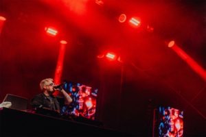 Nerds at Raves live DJ performt auf der Bühne des Elbenwald Festivals, umgeben von roten Lichtern und Nebel. Im Hintergrund sind visuelle Effekte auf großen Bildschirmen zu sehen, die die energiegeladene Atmosphäre des Events unterstreichen.