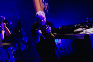 Nerds at Raves live DJ auf der Bühne des Elbenwald Festivals, umgeben von buntem Licht. Der Künstler zeigt eine provokante Geste mit den Fingern und hält ein Mikrofon in der Hand. Im Hintergrund ist das DJ-Equipment sichtbar, während ein Fotograf ihn festhält.