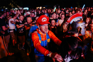 Nerds at Raves live Menschenmenge beim Elbenwald Festival, mit einem Besucher in einem Mario-Kostüm und einem anderen in einer römischen Rüstung. Die Atmosphäre ist lebhaft und bunt, während die Teilnehmer die Musik und das Gemeinschaftsgefühl genießen.
