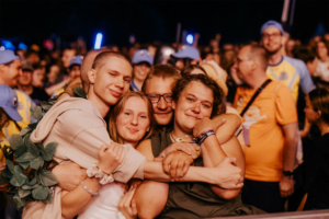 Nerds at Raves live Eine Gruppe von vier Freunden umarmt sich fröhlich inmitten der Menge beim Elbenwald Festival. Die Atmosphäre ist lebhaft, mit Menschen im Hintergrund, die die Musik und das Gemeinschaftsgefühl genießen. Ideal für Fans von Musik und Popkultur.