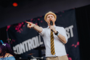 Olaf Unzel Ein Moderator auf der Bühne des Elbenwald Festivals, der das Publikum mit energiegeladener Ansprache begeistert. Im Hintergrund ist ein Banner mit dem Schriftzug "Kontrollfest" zu sehen. Die Atmosphäre ist lebhaft und festlich.