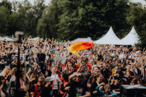 Eine große Menschenmenge feiert beim Elbenwald Festival, umgeben von Bäumen und Zelten. Die Teilnehmer tragen bunte Kostüme und zeigen Begeisterung, während sie mit Fahnen und Accessoires eine lebendige Atmosphäre schaffen.