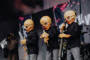 Drei Musiker in Alien-Masken spielen auf der Bühne beim Elbenwald Festival, umgeben von bunten Lichtern und einer festlichen Atmosphäre. Im Hintergrund ist ein Banner mit dem Festivalnamen sichtbar.