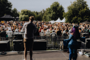Opening Blick von der Bühne beim Elbenwald Festival, mit einem Moderator und einer bunt gekleideten Performer im Vordergrund, während eine große Menge von Fans, darunter Stormtrooper-Kostüme, begeistert zuschaut. Musik und Cosplay vereinen sich in einer lebhaften Atmosphäre.