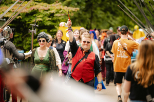 Bunte Parade von Festivalbesuchern beim Elbenwald Festival, gekleidet in fantasievolle Kostüme, während sie fröhlich durch den Park ziehen. Im Vordergrund sind ein lächelnder Mann in Weste und eine Frau in grüner Robe zu sehen, umgeben von weiteren Teilnehmern.