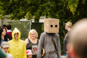 Eine Gruppe von Festivalbesuchern in kreativen Kostümen, darunter eine Person mit einer Papiertüte als Kopf und eine in einem gelben Regenmantel. Die Atmosphäre ist lebhaft und verspielt, perfekt für das Elbenwald Festival, das Musik und Nerd-Kultur vereint.