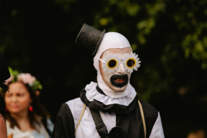Ein kostümierter Teilnehmer des Elbenwald Festivals trägt eine schwarze und weiße Verkleidung, ergänzt durch auffällige Sonnenbrillen mit Blumenmotiven. Im Hintergrund sind weitere Festivalbesucher zu sehen, die zur lebhaften Atmosphäre beitragen.
