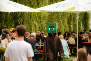 Person in einem Minecraft-Kostüm mit grünem Kopf und braunem Umhang, die durch eine belebte Festivalatmosphäre beim Elbenwald Festival geht, umgeben von Menschen in bunten Kostümen und festlicher Stimmung unter schattenspendenden Bäumen.