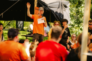 Percy Jackson Talk Ein Moderator in orange T-Shirt steht auf der Bühne des Elbenwald Festivals und spricht zu einem begeisterten Publikum. Im Vordergrund sind Teilnehmer in bunten Outfits zu sehen, die aktiv mitmachen und die Atmosphäre der Veranstaltung genießen.