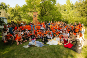 Percy Jackson Talk Gruppenfoto von Teilnehmern des Elbenwald Festivals, gekleidet in orangefarbene T-Shirts und Kostüme, umgeben von Bäumen und einem Banner. Die fröhliche Atmosphäre spiegelt die Mischung aus Musik und nerdiger Kultur wider.