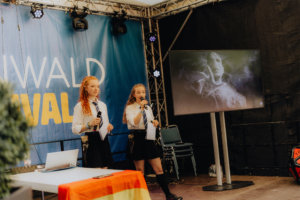 TypischRavenclaw Lesung Zwei Moderatorinnen stehen auf der Bühne des Elbenwald Festivals und halten Mikrofone. Im Hintergrund ist ein Bildschirm mit einem mystischen Bild zu sehen. Auf dem Tisch liegt ein Laptop und eine Flagge. Die Atmosphäre ist kreativ und lebhaft.