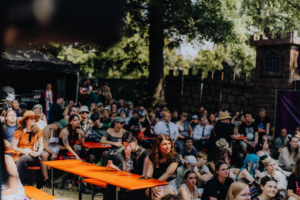 TypischRavenclaw Lesung Eine große Menschenmenge sitzt an roten Tischen unter Bäumen beim Elbenwald Festival. Die Teilnehmer, in bunten Outfits und mit verschiedenen Accessoires, genießen die Atmosphäre eines einzigartigen Musik- und Kulturereignisses.