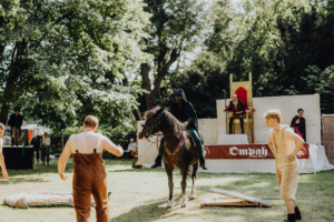 Ompah Kunstreitshow Ein Reiter in schwarzer Robe auf einem Pferd, umgeben von Darstellern in historischer Kleidung, während einer Aufführung auf dem Elbenwald Festival. Im Hintergrund sind Zuschauer und eine Bühne mit einem Thron zu sehen, die das mittelalterliche Ambiente unterstreichen.