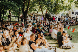 Ompah Kunstreitshow Eine große Menschenmenge sitzt auf dem Gras beim Elbenwald Festival, umgeben von Bäumen und festlicher Dekoration. Die Teilnehmer applaudieren und genießen die Atmosphäre eines einzigartigen Musik- und Nerd-Events unter freiem Himmel.