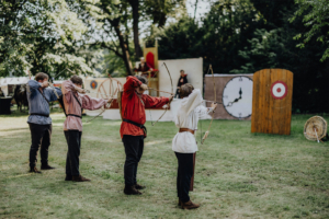 Ompah Kunstreitshow Vier Teilnehmer in historischen Kostümen üben das Bogenschießen auf dem Elbenwald Festival, umgeben von einer grünen Wiese und Zielscheiben im Hintergrund. Die Atmosphäre vereint Elemente von Musik und Fantasy.