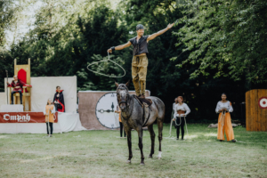 Ompah Kunstreitshow Ein Reiter steht auf einem grauen Pferd und jongliert mit einem Lasso während einer Aufführung auf dem Elbenwald Festival. Im Hintergrund sind Darsteller in Kostümen und Kulissen zu sehen, die die festliche Atmosphäre unterstreichen.