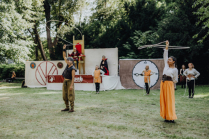 Ompah Kunstreitshow Darsteller in historischen Kostümen führen bei dem Elbenwald Festival eine beeindruckende Hula-Hoop-Show auf. Im Hintergrund sind Bühnenbilder und weitere Teilnehmer zu sehen, die die kreative Atmosphäre des Festivals unterstreichen.