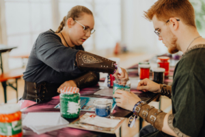 Retro-Basteln Zwei Teilnehmer des Elbenwald Festivals arbeiten an einem Basteltisch. Die junge Frau in einem mittelalterlichen Outfit füllt ein Glas mit buntem Material, während der junge Mann daneben konzentriert zuschaut. Bunte Gläser und Bastelmaterialien liegen auf dem Tisch.