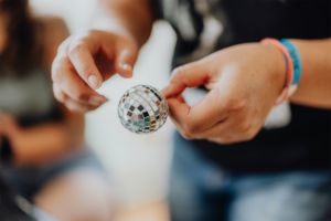 Retro-Basteln Hände halten eine kleine Discokugel auf dem Elbenwald Festival, umgeben von bunten Festivalarmbändern. Die Szene spiegelt die kreative und festliche Atmosphäre des Events wider, das Musik und Nerdkultur vereint.