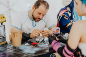 Retro-Basteln Ein Teilnehmer des Elbenwald Festivals arbeitet konzentriert an einem Bastelprojekt, umgeben von Materialien wie Scheren und Bastelelementen, während im Hintergrund eine Cosplayerin in aufwendigem Kostüm zu sehen ist.